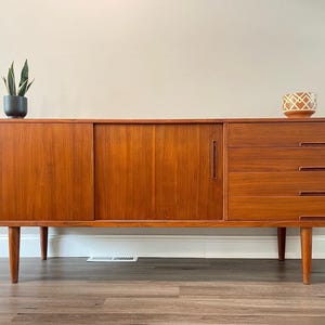 Op de afbeelding: Een lang houten dressoir met twee schuifdeuren met verticale verzonken handgrepen en een gedeelte aan de rechterkant met vier gestapelde lades. Het meubelstuk rust op vier taps toelopende poten en heeft een warme bruine houtnerfafwerking.