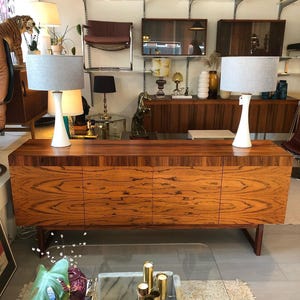 May include: A long wooden sideboard with a prominent grain pattern, featuring four cabinet doors and a dark wood top edge. Two white table lamps with light grey drum shades are positioned on the left and right sides of the sideboard surface.