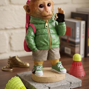 Könnte beinhalten: Eine Affenfigur trägt eine grüne Daunenjacke, beige Shorts sowie grün-weiße Sneaker. Der Affe hat einen pinken Rucksack, einen goldenen Ohrring und macht eine OK-Handgeste. Er steht auf einem runden Sockel auf einem grauen Steinblock.