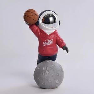 Könnte beinhalten: Eine Astronautenfigur in einem roten Kapuzenpullover mit dem aufgedruckten Wort GALAXY auf der Vorderseite. Der Astronaut hat ein reflektierendes Helmvisier, hält einen braunen Basketball über dem Kopf und steht auf einer grauen Mondbasis.