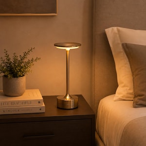 Peut inclure: Une lampe de table métallique, haute et élancée, dotée d'une base circulaire et d'un sommet plat en forme de disque qui émet une lueur chaude dirigée vers le bas. La lampe présente une finition brossée lisse et un pied vertical au design épuré.