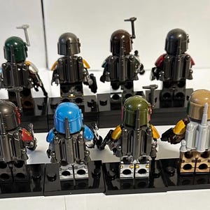 May include: Eight plastic minifigures standing on black rectangular display bases. Each figure wears a helmet and a jetpack, featuring various colour schemes including green, blue, tan, and dark grey, viewed from the back in two rows.