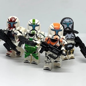 May include: Four plastic toy soldier minifigures standing in a row. Each figure wears a distinct helmet and armor set featuring unique color patterns in red, green, orange, and black. Each figure holds a black plastic toy blaster rifle accessory.