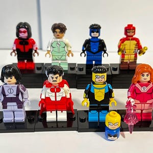 Puede incluir: Una colección de ocho minifiguras de bloques de construcción de plástico con varios personajes en coloridos trajes de superhéroes, incluyendo atuendos rojos, azules, amarillos y morados, cada una sobre una base cuadrada negra con una cabeza extra.