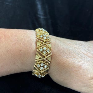 May include: A gold-tone metal bracelet featuring a repeating geometric pattern of diamond-shaped links. Each link is textured with fine lines and contains a cluster of small, clear, sparkling stones set in the center of the diamond shapes.