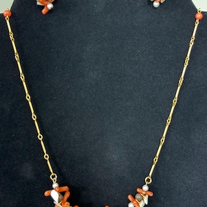 Puede incluir: Un conjunto de collar de cadena en tono dorado y pendientes de clip a juego. El collar presenta una cadena delicada acentuada con racimos de ramas de color naranja tipo coral, pequeñas perlas blancas y cuentas de color marrón oscuro.