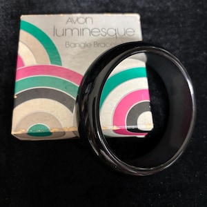 Peut inclure: Un bracelet jonc noir brillant placé devant sa boîte en carton carrée d'origine. La boîte porte l'inscription "AVON luminesque Bangle Bracelet" et un motif graphique de cercles concentriques dans des tons de vert, rose, noir et beige.