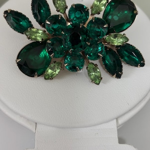 Puede incluir: Un broche que presenta un conjunto de piedras de vidrio facetadas en color verde esmeralda y verde lima claro, engastadas en una montura de metal. El diseño incluye formas redondas, de pera y marquesa dispuestas en un patrón floral con garras.