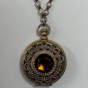 May include: A round gold-tone locket pendant suspended from a matching chain. The locket features an intricate filigree border surrounding a large, faceted, dark amber-colored gemstone set in the center. The metal has a polished, metallic finish.
