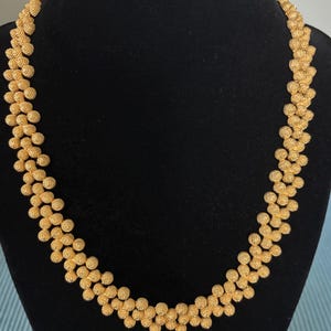 Può includere: Una collana color oro caratterizzata da un motivo ripetitivo di piccole perle sferiche testurizzate disposte in una catena densa e interconnessa. La superficie metallica delle perle presenta una finitura finemente granulata che cattura la luce.