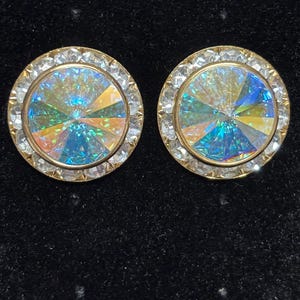 May include: A pair of round stud earrings featuring a large, faceted, iridescent central stone that displays shifting blue, green, and yellow hues. The central stone is surrounded by a circular border of small, clear, white crystals set in gold-toned metal.