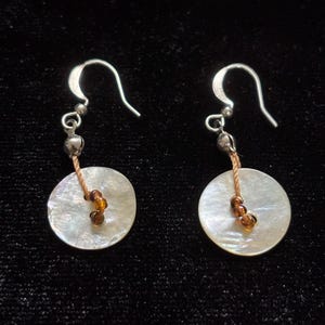 May include: A pair of drop earrings featuring circular white mother-of-pearl discs suspended from silver-toned metal hooks. Each earring includes a small silver-toned bead, a tan cord, and a single amber-colored glass bead at the center of the disc.