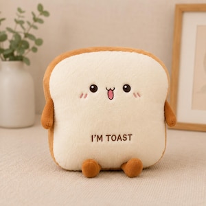 May include: A plush toy shaped like a slice of bread with a light cream-coloured centre and brown crust edges. It features a stitched face with black eyes, a pink tongue, and the brown embroidered text I'M TOAST, along with small brown arms and feet.