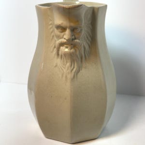 May include: A beige ceramic pitcher featuring a molded relief of a bearded face on the front. The vessel has a hexagonal body shape, a wide pouring spout, and a handle extending from the back. The surface has a smooth, glazed finish.