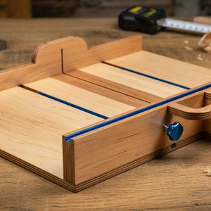 Peut inclure: Un traîneau de défonceuse en bois clair doté d'une base plate, d'un guide perpendiculaire et d'un mécanisme coulissant avec un bouton bleu et des inserts en T bleus. Une étiquette en papier blanc porte l'inscription Custom Router Sled.
