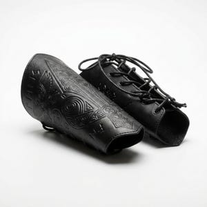 May include: A pair of black leather bracers featuring intricate embossed designs of a bird and geometric patterns, along with runic symbols. The bracers include black cord lacing through metal eyelets for a secure fit around the forearm.