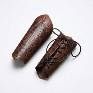 May include: A pair of brown arm bracers featuring embossed intricate patterns, including a central sword motif and runic symbols. One bracer is flat, while the other is curved and secured with black cord lacing through metal eyelets.