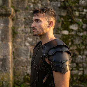 May include: A black leather chest and shoulder armor piece featuring layered pauldrons with scalloped edges. The armor is constructed with multiple panels secured by metal rivets and includes adjustable leather straps along the side for a secure fit.