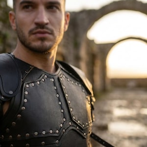May include: A black leather chest armor piece featuring structured panels and metallic silver studs along the seams. The armor is constructed with multiple overlapping sections and reinforced stitching, providing a rigid and protective appearance.