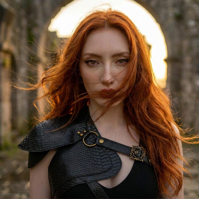 May include: A black textured shoulder armor piece featuring a snakeskin-like pattern, secured with a brass-toned metal ring and a decorative ornate buckle. The armor is worn over a simple black top, highlighting the contrast between the dark material.