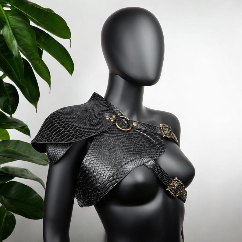 May include: A black shoulder armor piece featuring a textured snakeskin pattern, secured with brass-toned metal rings and ornate rectangular buckles. The structured garment covers one shoulder and extends across the chest with adjustable straps.