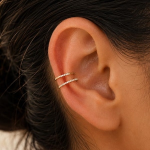 May include: A gold ear cuff featuring two thin, parallel bands. Each band is encrusted with a row of small, clear, sparkling stones that catch the light. The jewelry is positioned on the upper cartilage of the ear, creating a layered appearance.