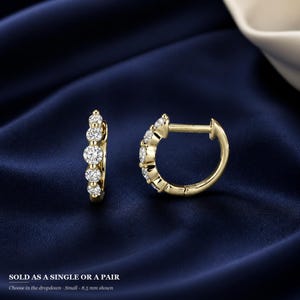 May include: A pair of gold hoop earrings, each featuring five round-cut clear stones set along the front curve. The earrings have a hinged closure mechanism. Text at the bottom reads: SOLD AS A SINGLE OR A PAIR. Choose in the dropdown - Small - 8.5 mm shown.