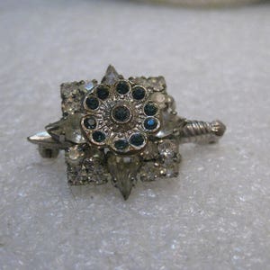 May include: A silver tone brooch with a blue rhinestone center surrounded by clear rhinestones. The brooch is shaped like a star with a sword design.