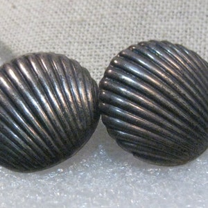 May include: A pair of silver tone shell shaped earrings. The earrings are textured with lines that resemble the ridges of a seashell.