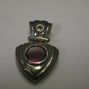 May include: Silver pendant with a heart-shaped design and a red gemstone in the centre.
