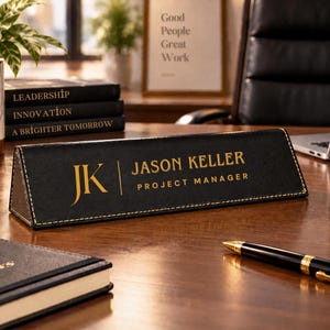 May include: A black triangular desk nameplate with gold stitching and gold lettering that reads JK, JASON KELLER, and PROJECT MANAGER. The nameplate is positioned on a wooden surface alongside a black notebook, a black pen, and a black mug.