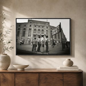 May include: A black and white framed photograph of the exterior of Yankee Stadium. Several individuals stand near a sign that reads TO GATES 3-4-5. The architecture features large arched windows and stone detailing, capturing a historic scene.
