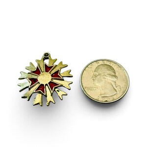 May include: A gold-toned metal Maltese cross pendant featuring red enamel accents on the arms and a central circular medallion. The pendant is positioned next to a standard United States quarter coin, which measures approximately 0.955 inches in diameter.