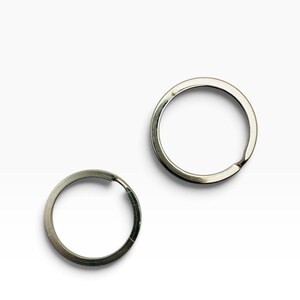 May include: Two circular silver-toned metal split rings for keychains. Each ring features a flat, coiled wire design with a small overlapping end section, providing a secure loop for attaching keys or other small items to a keyring assembly.
