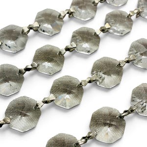 May include: A decorative garland consisting of multiple strands of clear, faceted, octagonal glass crystals. Each crystal is connected to the next by small, metallic, fan-shaped silver-toned connectors, creating a continuous, reflective chain.