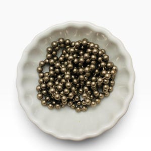 May include: A coiled metallic ball chain with a dark silver finish resting inside a small, white, scalloped-edge ceramic bowl. The chain consists of numerous tiny, interconnected spherical beads that create a textured, reflective surface.