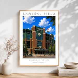 May include: A framed vertical wall art print featuring Lambeau Field, the home of the Green Bay Packers. The image shows the stadium's brick facade, a clock, and flags under a blue sky. Text includes Lambeau Field, Green Bay Packers, and coordinates.