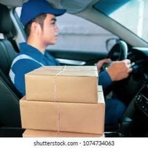 Pode incluir: Uma pilha de três caixas de envio de papelão marrom simples posicionadas em primeiro plano. As caixas são retangulares com costuras visíveis e empilhadas verticalmente. O fundo mostra o interior de um veículo com um motorista de uniforme.