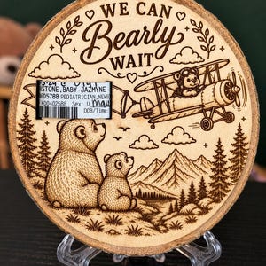 May include: A circular wood slice with a laser-engraved design featuring two bears looking at a biplane, mountains, and trees. The text reads WE CAN Bearly WAIT. A rectangular hospital identification band is attached to the left side of the wood.