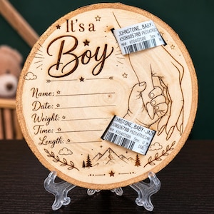 May include: A circular wooden plaque featuring laser-engraved text that reads It's a Boy, along with fields for Name, Date, Weight, Time, and Length. The design includes an illustration of an adult hand holding a baby hand with two hospital ID bands.