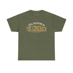 Könnte beinhalten: Ein olivgrünes Kurzarm-T-Shirt mit dem Text "STILL BELIEVING IN THE COMEBACK" in einer abgenutzten, goldenen Schriftart auf der Brust, einer dünnen horizontalen Linie unter dem Text und einem kleinen Logo in der Nähe des Halsausschnitts.