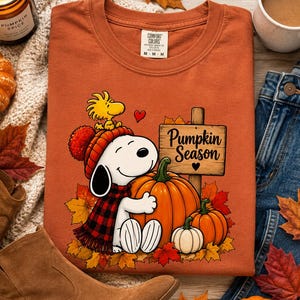 May include: A burnt orange t-shirt featuring a graphic of a white dog wearing a red plaid scarf and a red knit hat, hugging a large orange pumpkin. Next to the dog is a yellow bird and a wooden sign with the text Pumpkin Season in black.