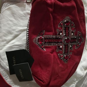 Puede incluir: Un bolso de mano de ante rojo con un dise&ntilde;o de cruz grande adornado con cristales rojos y transparentes. El bolso incluye una correa de cadena plateada y dos etiquetas negras con el texto VERSACE impreso en dorado sobre tela blanca.