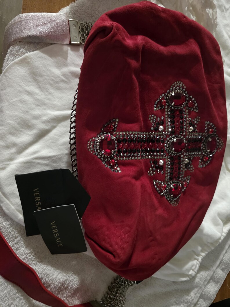 Puede incluir: Un bolso de mano de ante rojo con un dise&ntilde;o de cruz grande adornado con cristales rojos y transparentes. El bolso incluye una correa de cadena plateada y dos etiquetas negras con el texto VERSACE impreso en dorado sobre tela blanca.
