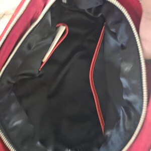 Puede incluir: El interior de un bolso rojo con un forro negro liso. El bolso tiene una cremallera plateada a lo largo del borde superior e incluye un peque&ntilde;o bolsillo abierto en el lateral con un borde rojo, ofreciendo una vista clara del compartimento.