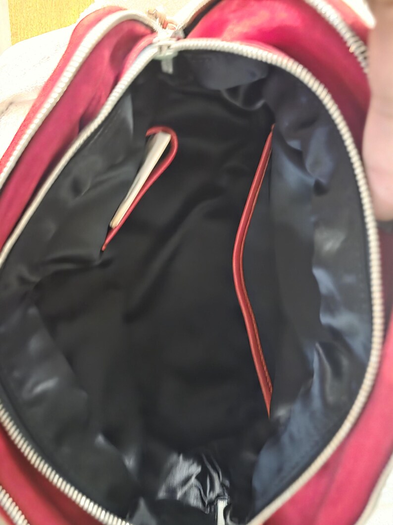 Puede incluir: El interior de un bolso rojo con un forro negro liso. El bolso tiene una cremallera plateada a lo largo del borde superior e incluye un peque&ntilde;o bolsillo abierto en el lateral con un borde rojo, ofreciendo una vista clara del compartimento.