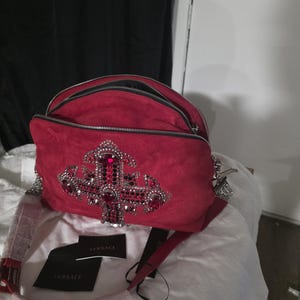 Puede incluir: Un bolso de hombro de ante rojo con un gran emblema de cruz adornado con cristales rojos y plateados. El bolso cuenta con dos compartimentos con cremallera, una correa roja a juego y dos etiquetas negras de la marca Versace con texto blanco.