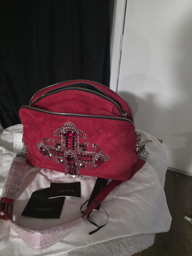 Puede incluir: Un bolso de hombro de ante rojo con un gran emblema de cruz adornado con cristales rojos y plateados. El bolso cuenta con dos compartimentos con cremallera, una correa roja a juego y dos etiquetas negras de la marca Versace con texto blanco.