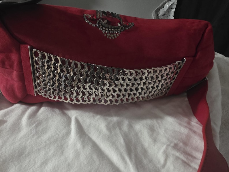Puede incluir: Un bolso de mano rojo que presenta un panel de malla met&aacute;lica plateada con un patr&oacute;n geom&eacute;trico en la parte frontal. El bolso est&aacute; adornado con un grupo decorativo de cristales transparentes y una piedra roja central sobre la malla met&aacute;lica.
