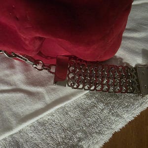Puede incluir: Un bolso de mano rojo brillante con una superficie texturizada y una correa de cadena de metal plateado. La correa presenta un patr&oacute;n de eslabones geom&eacute;tricos repetitivos y se conecta al bolso mediante un cierre de metal plateado.